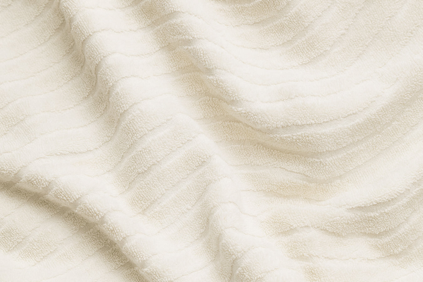 A close-up of a soft, cream-colored fabric with a subtle wave-like texture. The folds and shadows highlight its cozy and luxurious quality.