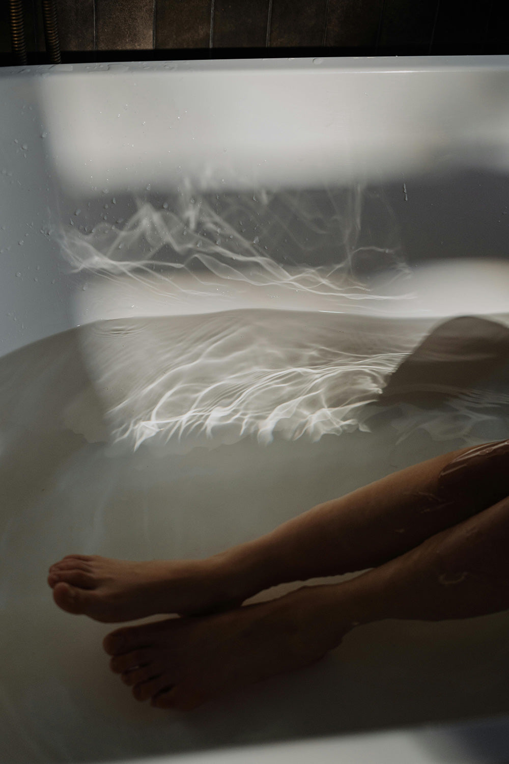 A person’s legs are partially submerged in bathwater with light reflections dancing on the surface. The image focuses on the calm water and soft lighting, creating a serene atmosphere.