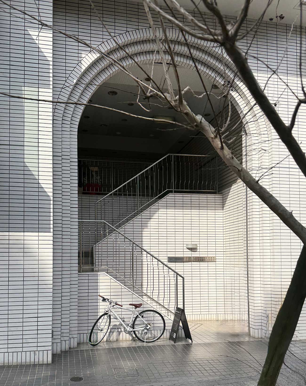 A white tiled building exterior with an arched entrance and a staircase leading upward. A white bicycle is parked near the stairs beside a black standing sign, with tree branches visible in the foreground.