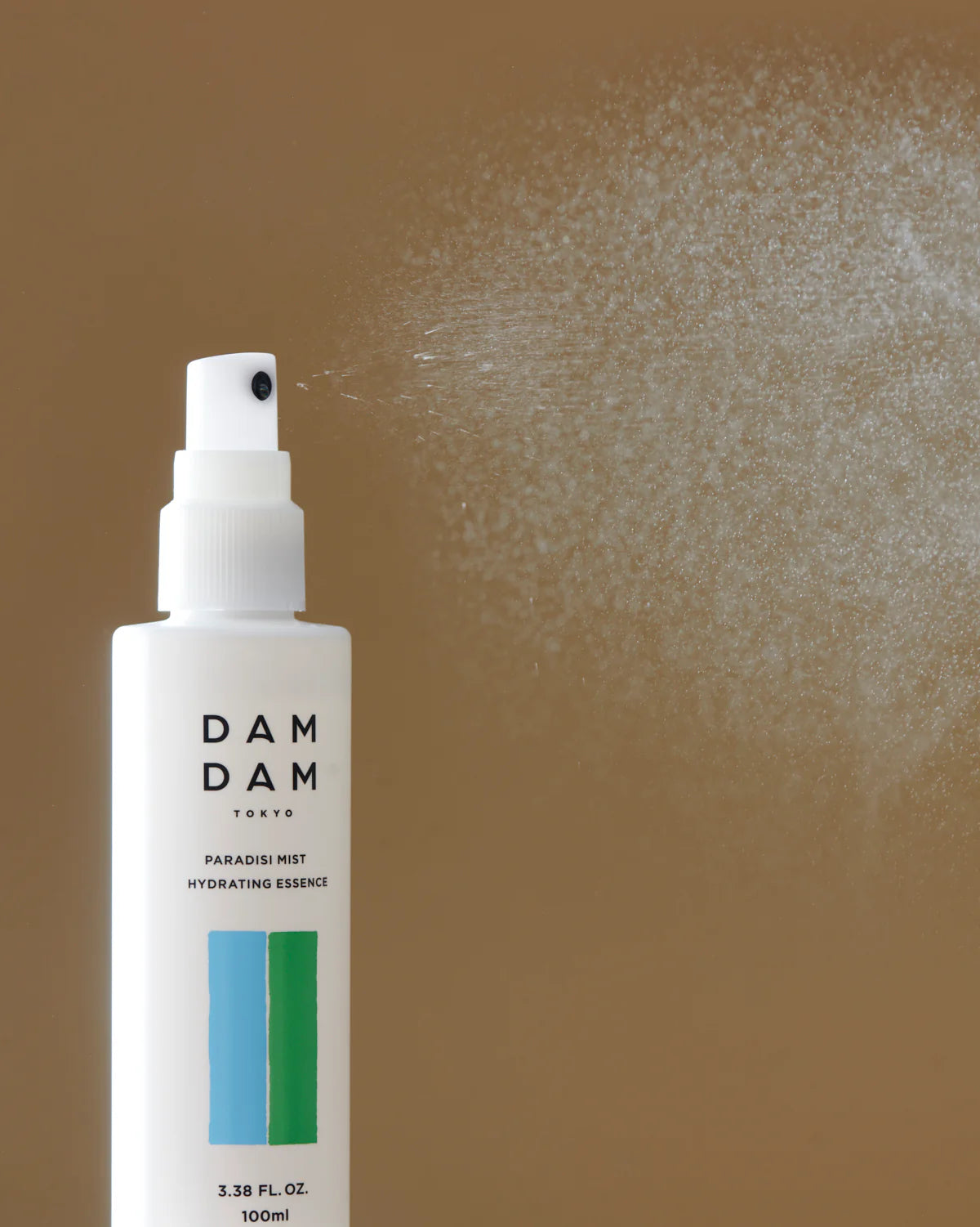 A bottle of DAMDAM Tokyo Paradisi Mist Hydrating Essence sprays a fine mist against a warm, neutral background. The image captures the refreshing, lightweight texture of the essence while emphasizing the clean, minimalist design of the packaging.