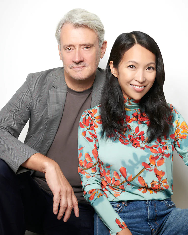A portrait of two people sitting closely together against a light background. They both appear relaxed and smiling, dressed in casual yet stylish clothing.