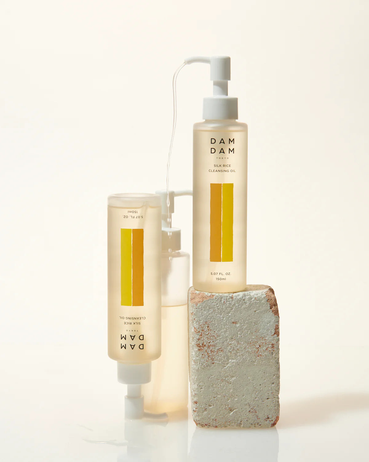 Bottles of DAMDAM Tokyo Silk Rice Cleansing Oil are arranged in a minimal still-life composition, with one upright bottle placed on a textured stone block. The golden liquid and gentle lighting highlight the oil’s purity and luxurious skincare appeal.