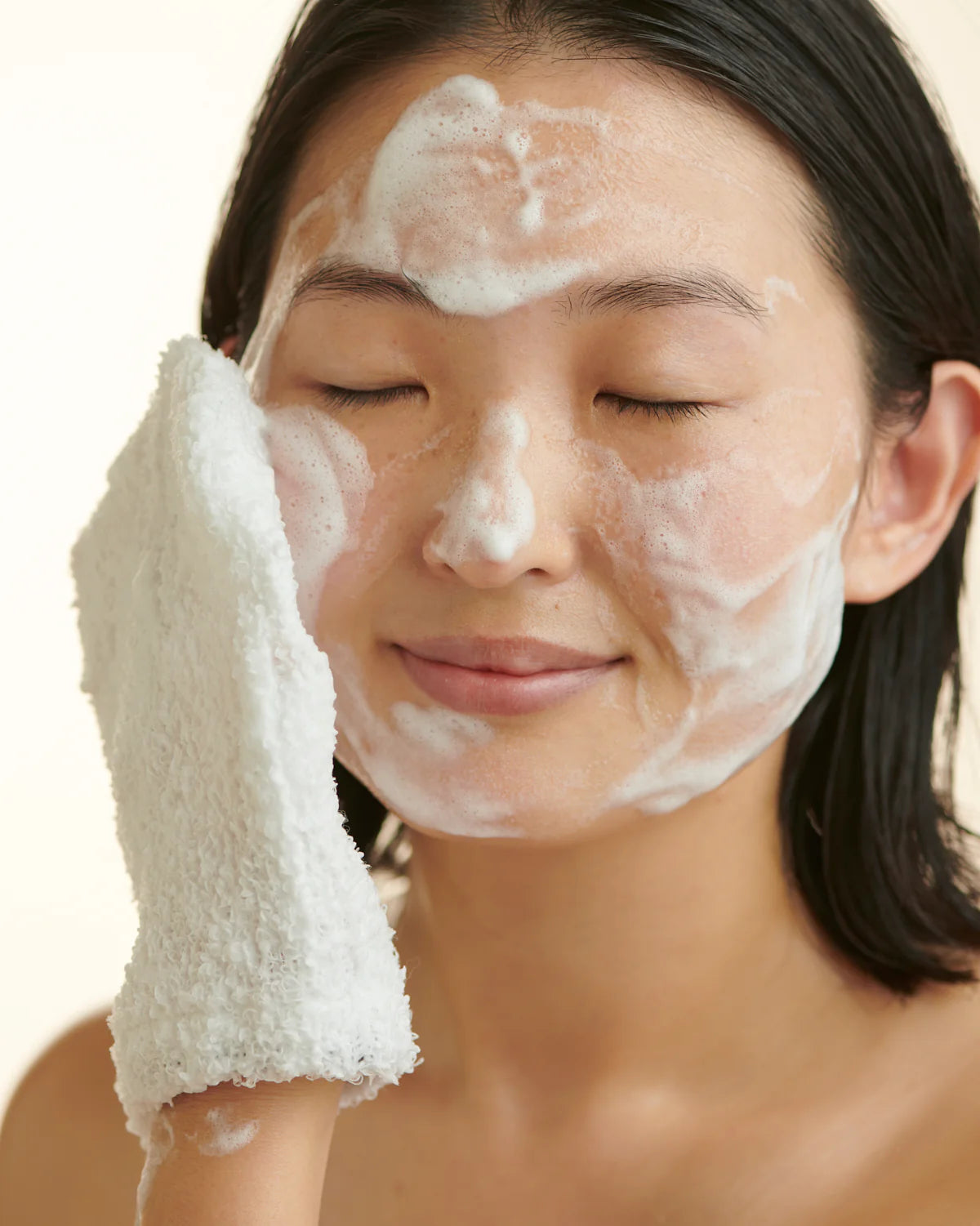 A person gently cleanses their face with a foaming cleanser, eyes closed and a soft towel pressed against their skin. The image conveys freshness, calm, and the soothing ritual of daily skincare.