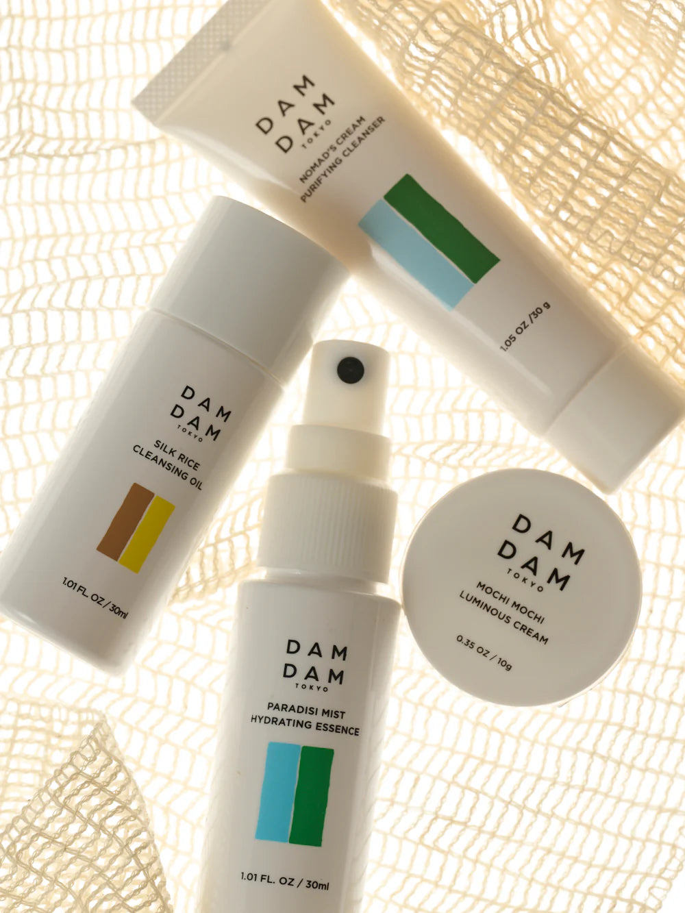 A flatlay of DAMDAM Tokyo skincare products, including a cleanser, cleansing oil, hydrating essence, and cream, arranged on a textured fabric surface. The warm lighting highlights the clean packaging and minimalist design.