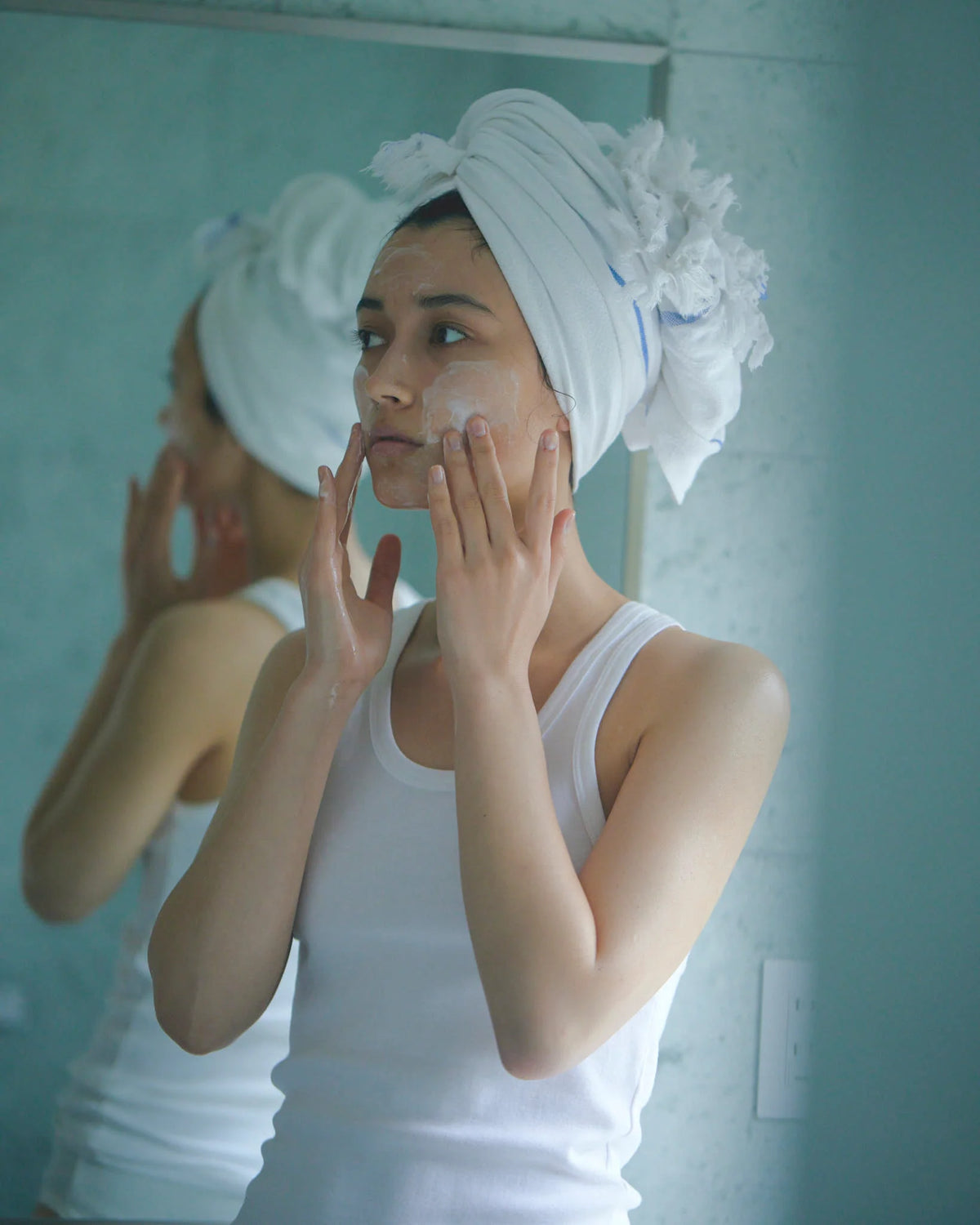 A person with a towel wrapped around their head applies facial cleanser in front of a bathroom mirror. The soft lighting and calm expression create a feeling of relaxation and self-care.