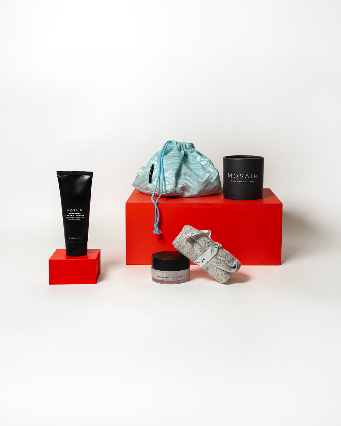 A curated display of Mosaik skincare items arranged on bold red blocks, featuring a light blue drawstring bag, a black product tube, a small jar, and a neatly rolled towel.