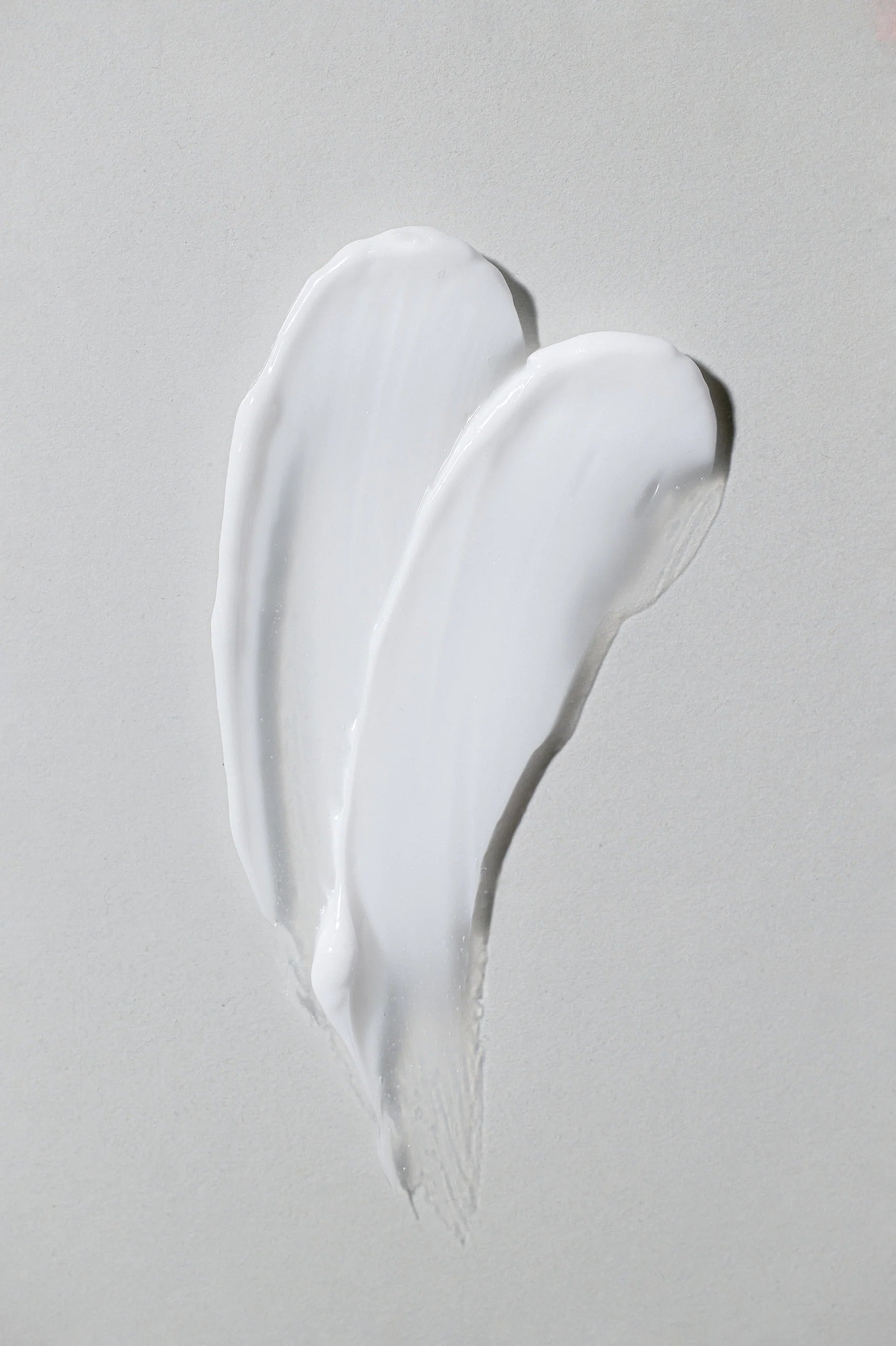 Close-up of two smooth white cream swipes on a light grey surface, showing the texture and consistency of a skincare product.