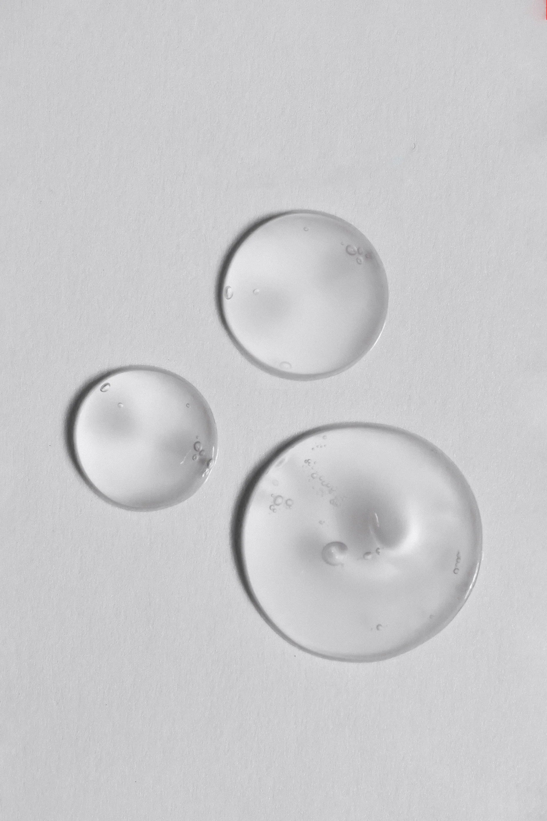 Close-up of three transparent gel drops on a white surface, showing smooth texture and light reflections, representing skincare serum or hydrating essence.