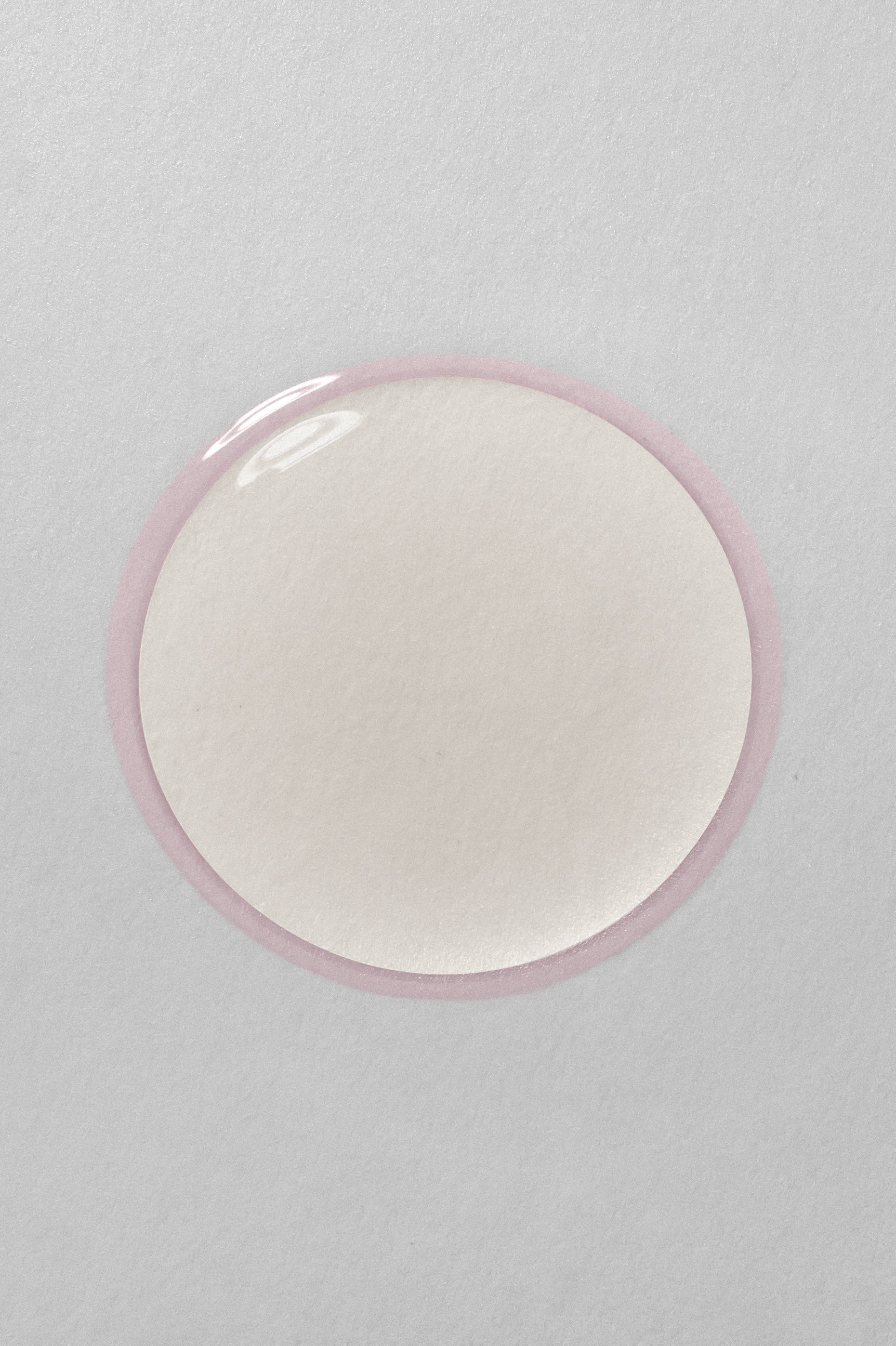 Close-up of a translucent liquid droplet with a soft pink edge on a smooth, light grey surface, capturing the texture and clarity of a skincare serum or essence.