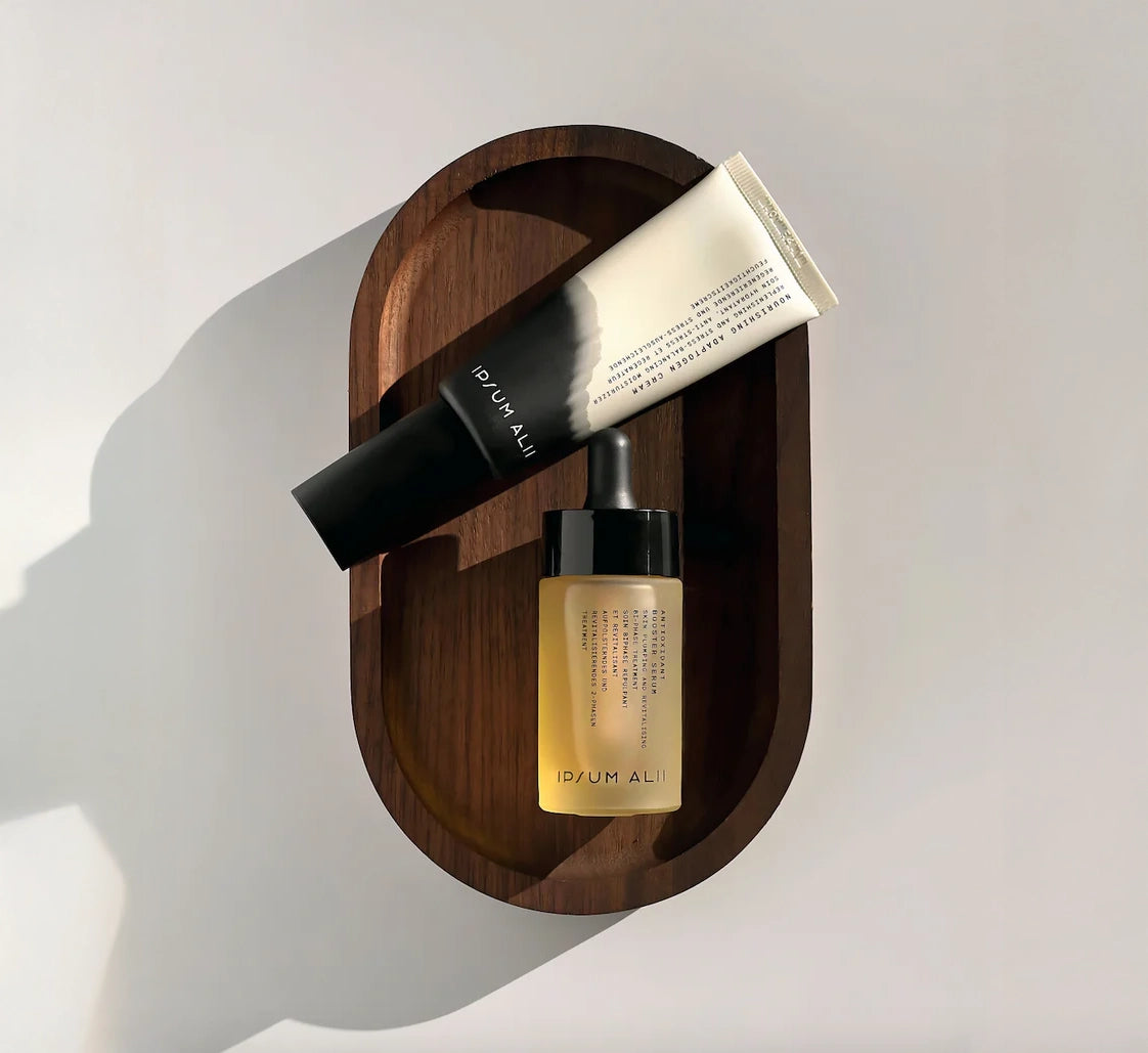 Two Ipsum Alii skincare products — a tube and a dropper bottle — rest neatly on a wooden tray against a soft, sunlit background. The minimalist packaging, featuring black gradients and clean typography, reflects a refined and modern aesthetic.