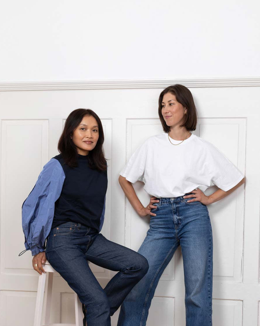Two women posing in a minimal, bright room with wooden floors and white panel walls, dressed in casual chic denim outfits — one sitting on a stool wearing a navy and blue top, and the other standing in a white t-shirt and red heels.