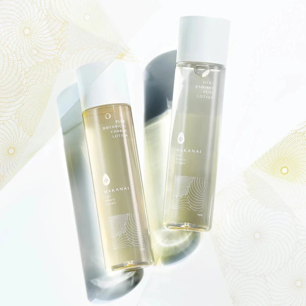 Two elegant Makanai lotion bottles are displayed upright on a reflective surface with soft golden patterns surrounding them.
