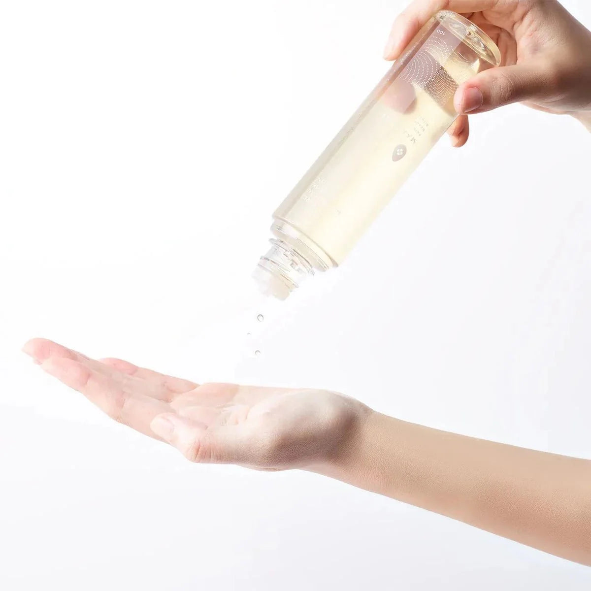 A hand tilts a clear bottle of lotion, letting droplets fall gently into an open palm against a bright white background.