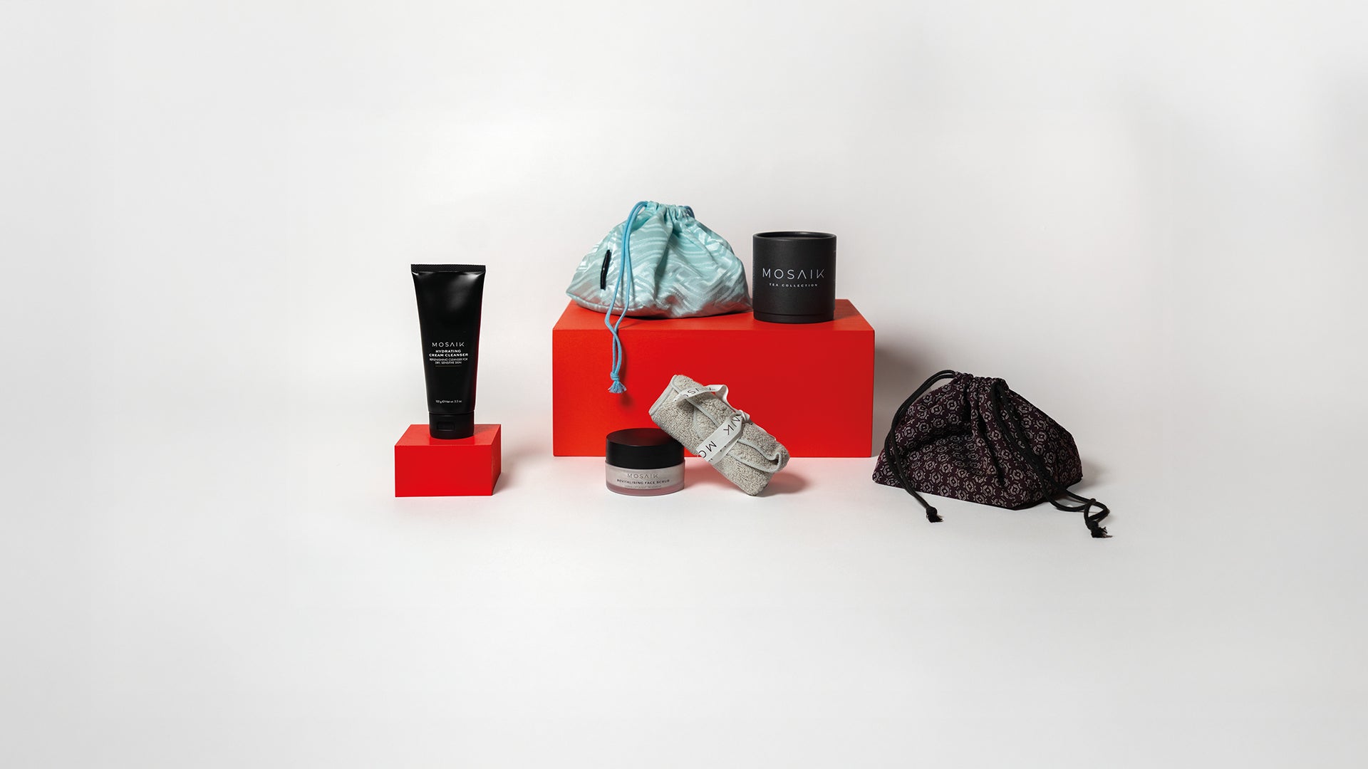 A curated display of Mosaik skincare items arranged on bold red blocks, featuring a light blue and brown  drawstring bag, a black product tube, a small jar, and a neatly rolled towel.