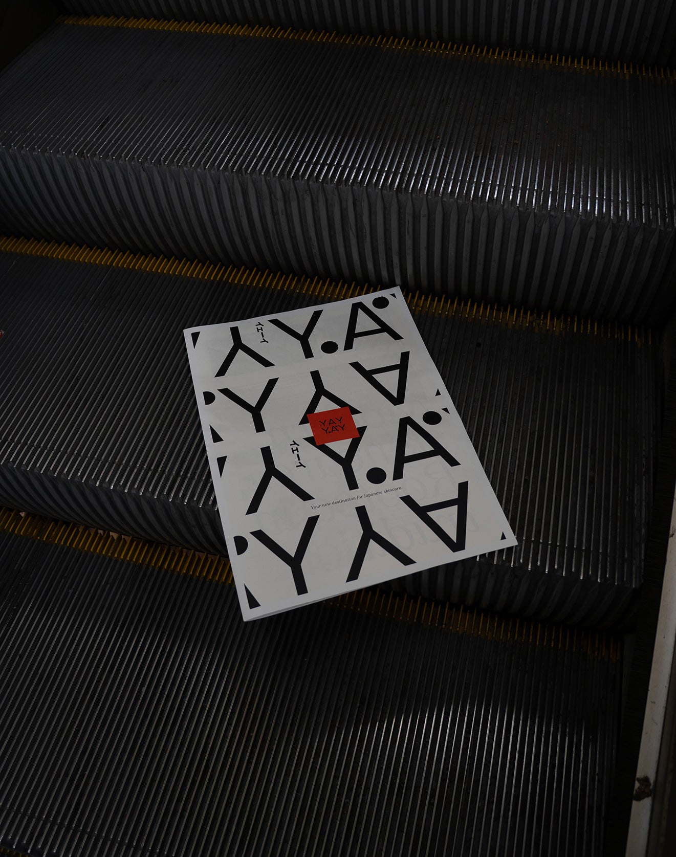 A printed sheet with the word “YAY” in a bold black pattern and a red hexagon logo placed on an escalator step. The metallic surface of the escalator contrasts with the white background of the paper.