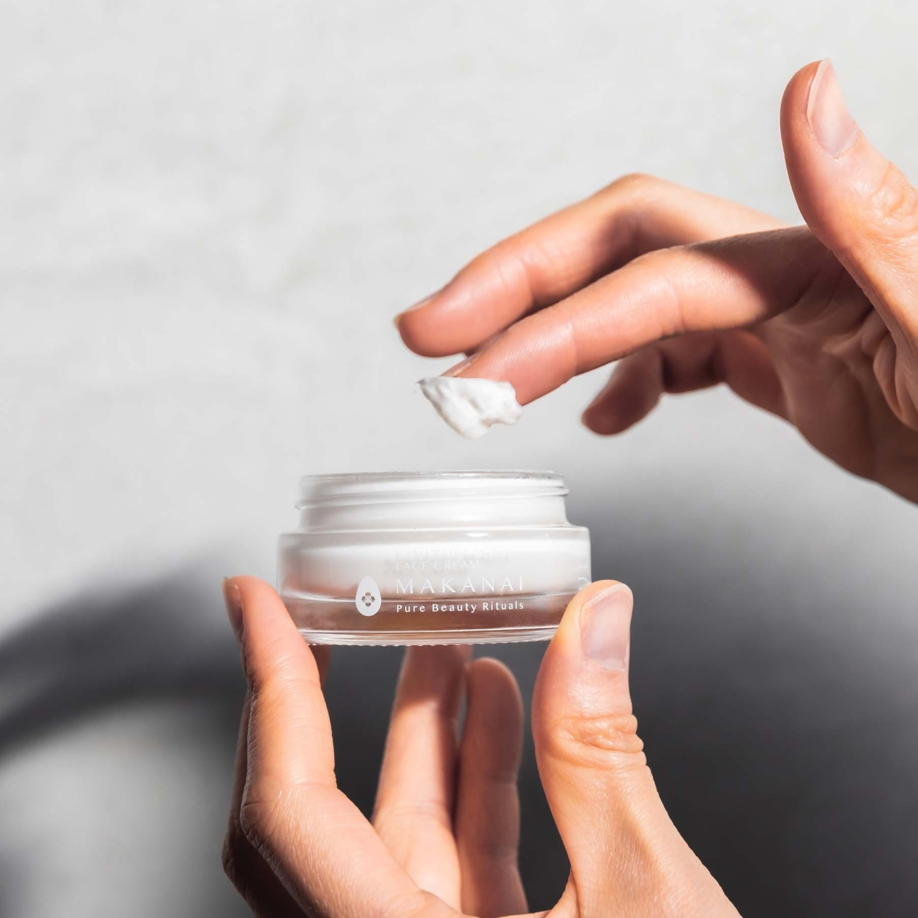 A person’s hands holding an open jar of Makanai Face Cream with a small amount of product on their fingertip. The jar is clear with white cream inside and minimal branding on the front.
