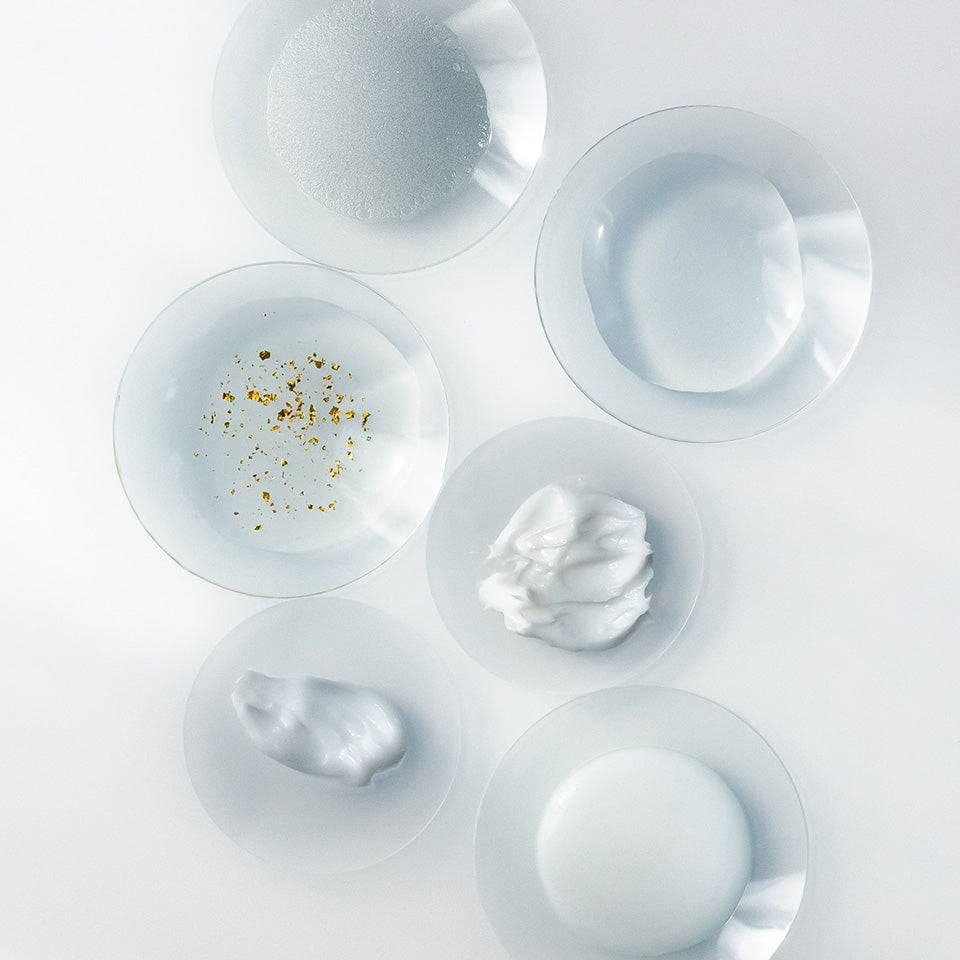 Six small glass dishes showing different skincare textures and consistencies, including creams, gels, foam, and a serum with gold flakes. The products are displayed on a white surface in soft, even lighting.