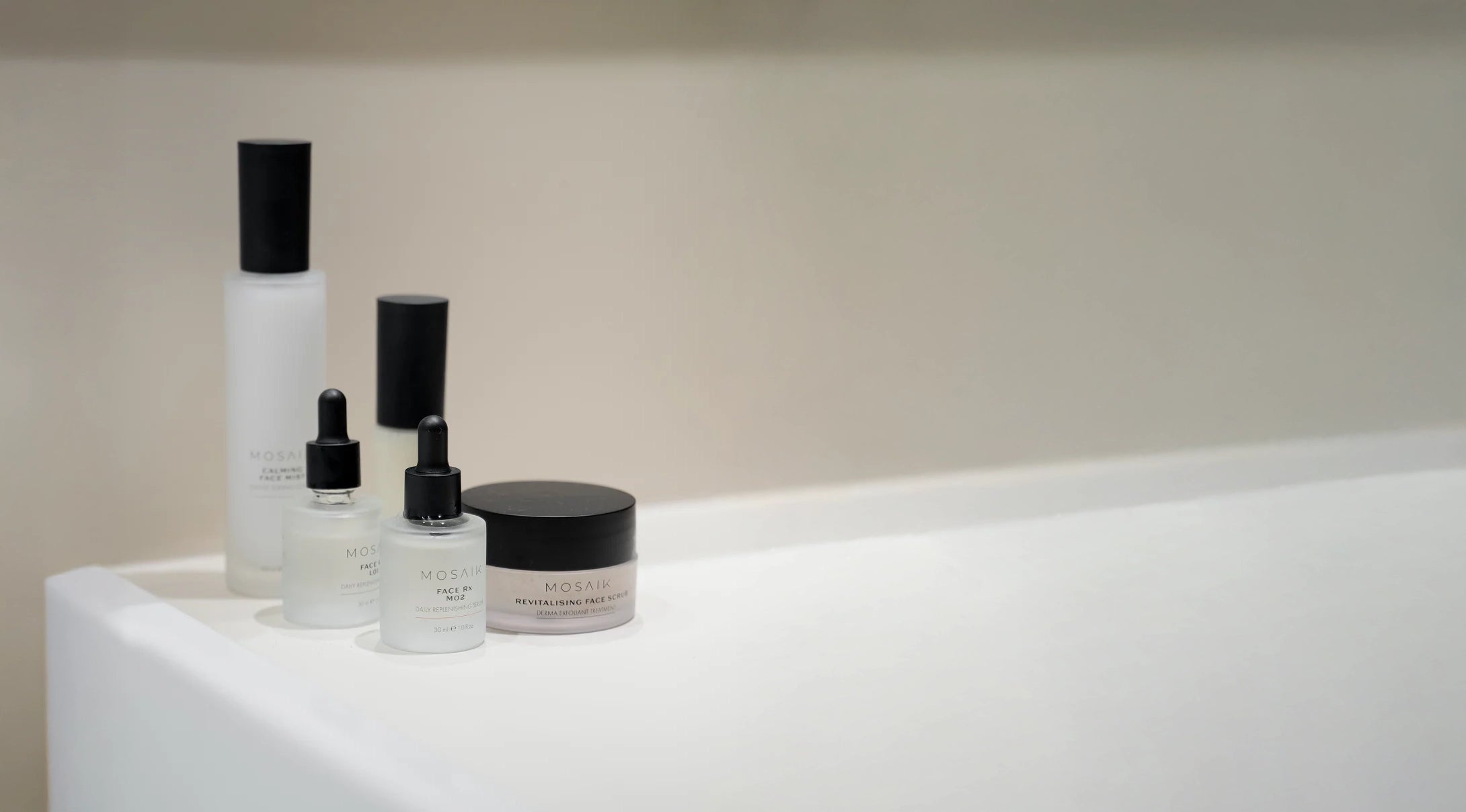 A minimalist arrangement of Mosaik skincare products is displayed on a clean white countertop against a soft beige background. The image conveys a calm, refined aesthetic emphasizing purity and modern simplicity.