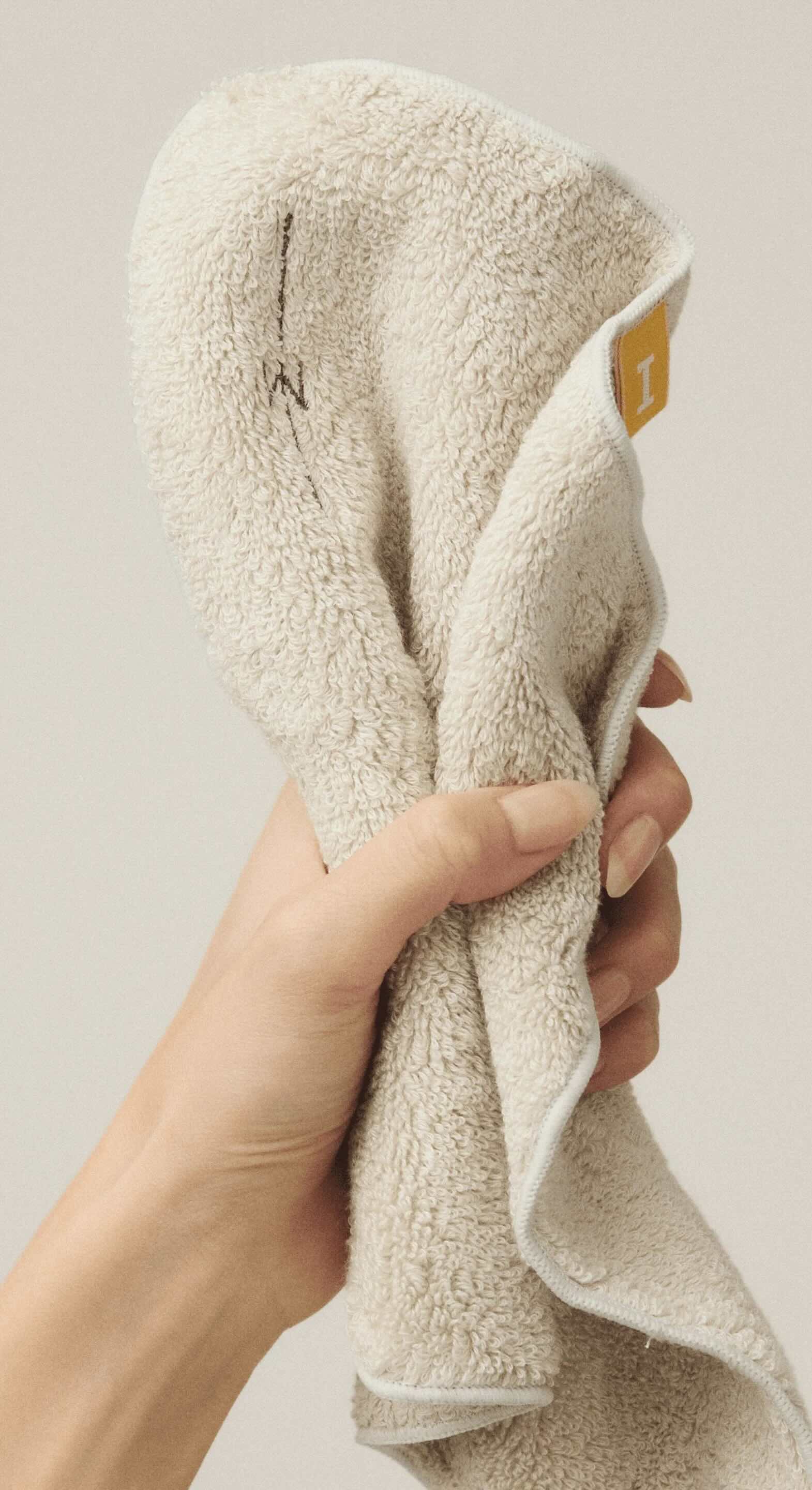 A hand gently holds a soft beige towel with a small yellow tag visible near the edge. The close-up image emphasizes texture, warmth, and everyday simplicity in skincare or self-care routines.