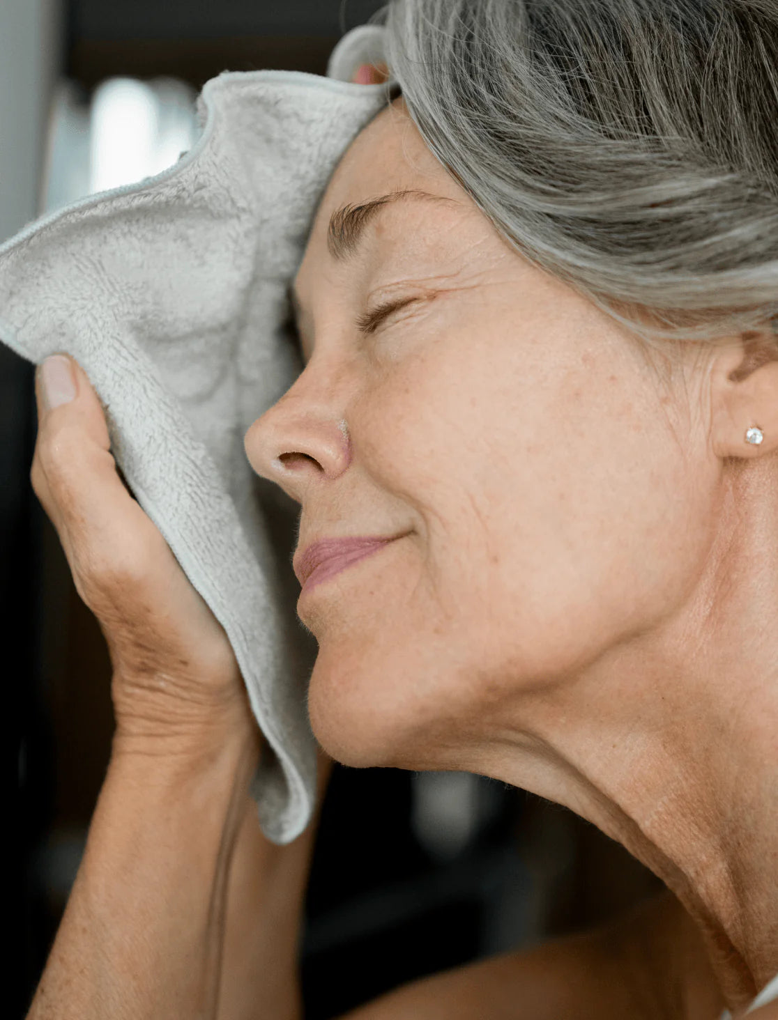 A mature woman with gray hair gently presses a soft towel against her face, eyes closed in relaxation. The image captures a calm, self-care moment emphasizing gentle skincare and well-being.