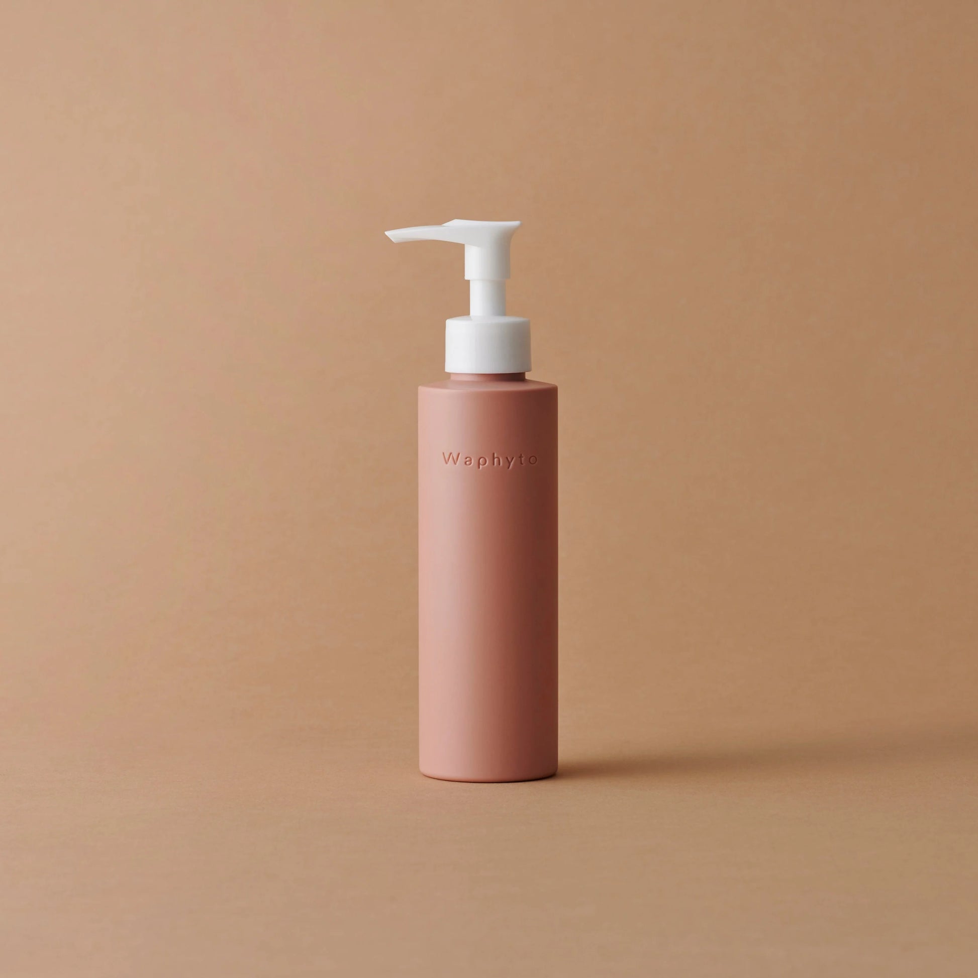 A peach-toned Waphyto bottle with a white pump sits on a neutral beige background. The minimalist composition highlights the clean and elegant packaging design.