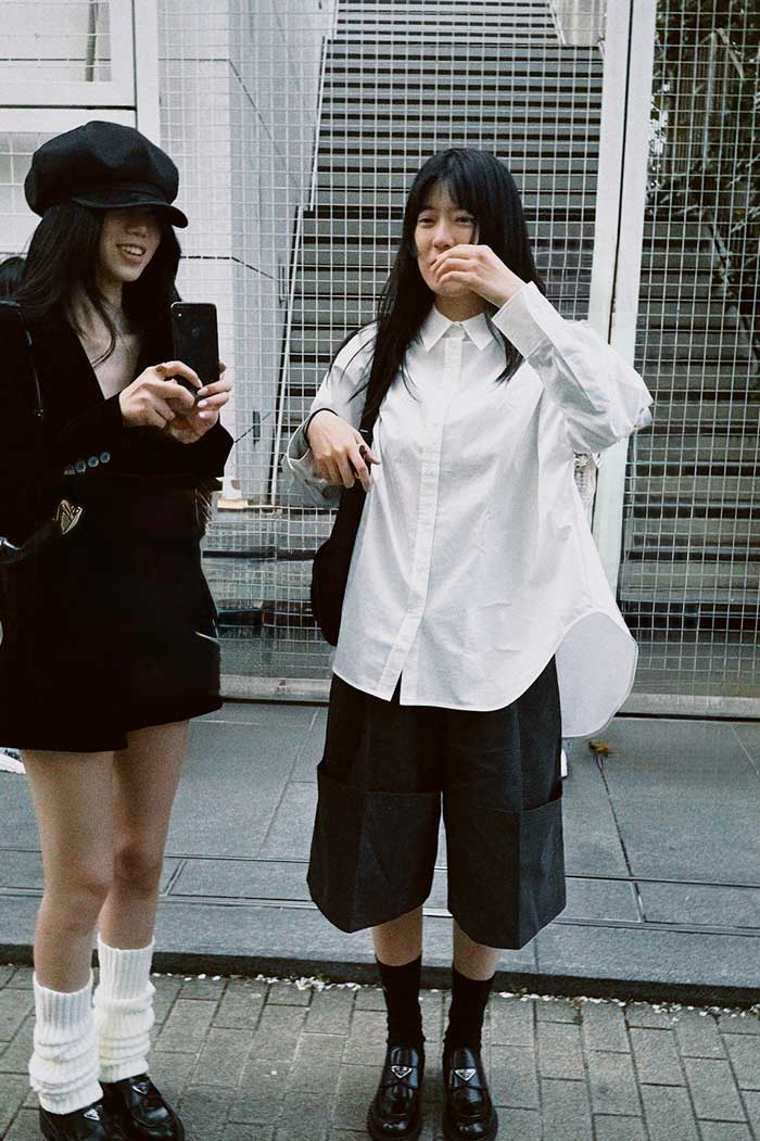 Two people standing outdoors in front of a staircase, one wearing a black outfit with a hat and the other in a white shirt and dark shorts. One person is taking a photo with a phone while the other appears to be laughing or talking.