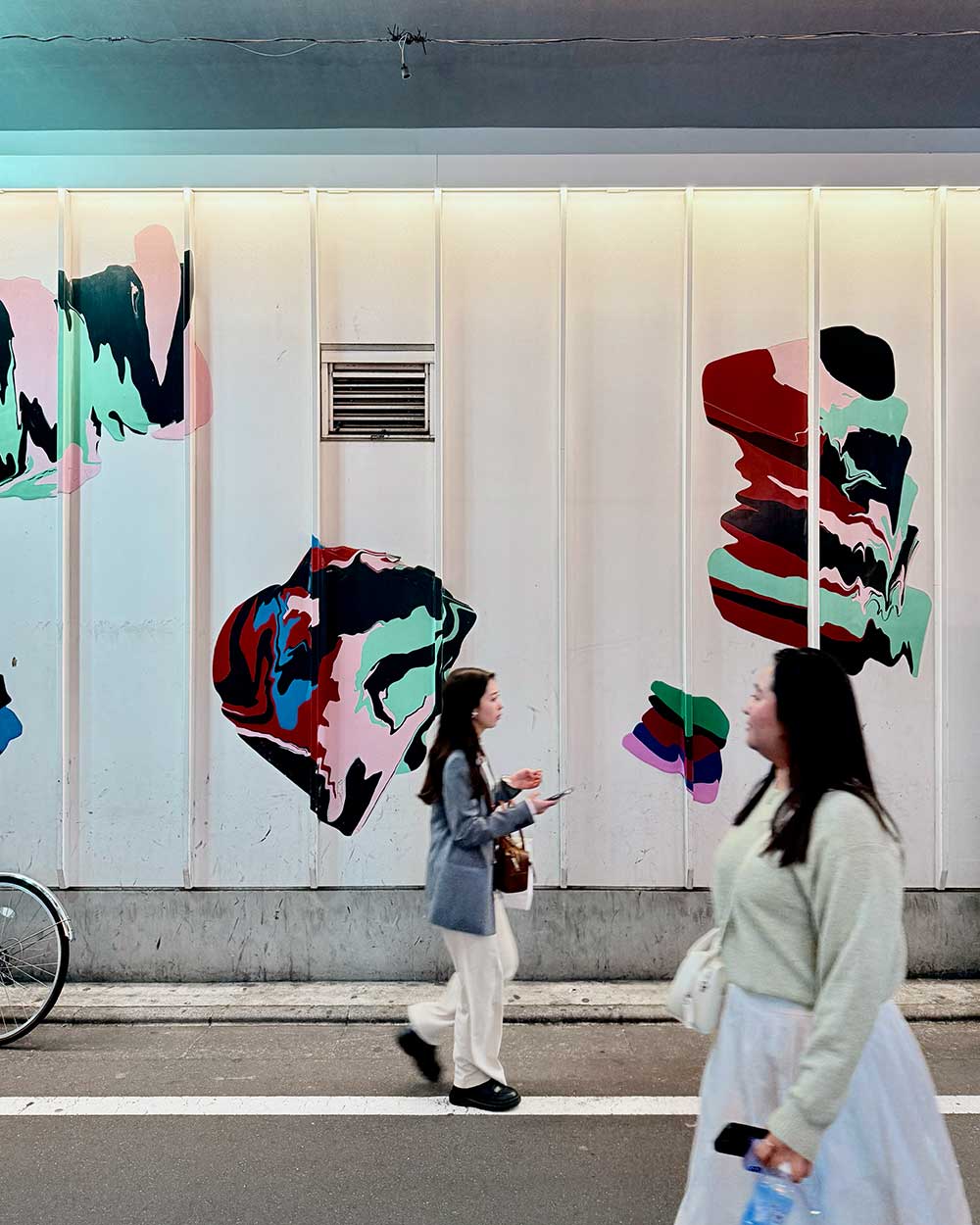 Two women walking past a white wall decorated with colorful abstract murals in red, green, black, and pink.
