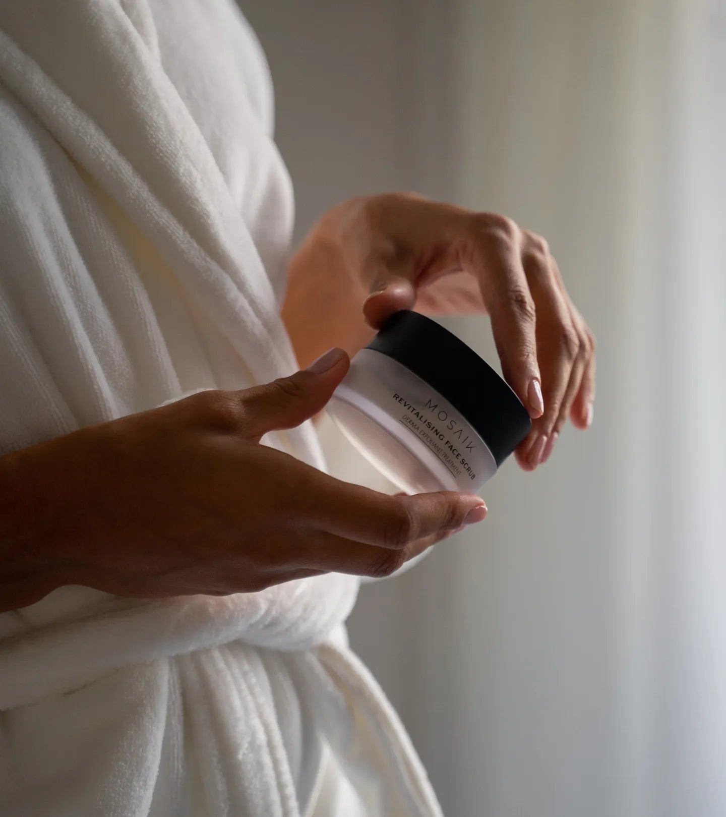 A person in a white robe holds a jar of Mosaik Revitalising Face Scrub in soft, natural light. The image conveys a calm, spa-like atmosphere emphasizing self-care and minimalist luxury.