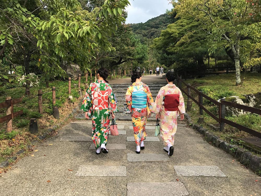 Three people wearing colorful kimonos walk side by side along a stone path in a lush, green park.