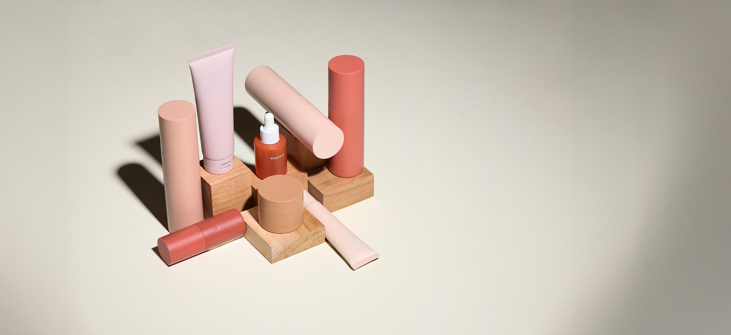 A collection of Waphyto skincare products in pink and terracotta-toned packaging displayed on wooden blocks. The arrangement includes tubes, jars, and a dropper bottle, shown on a neutral background with soft lighting.