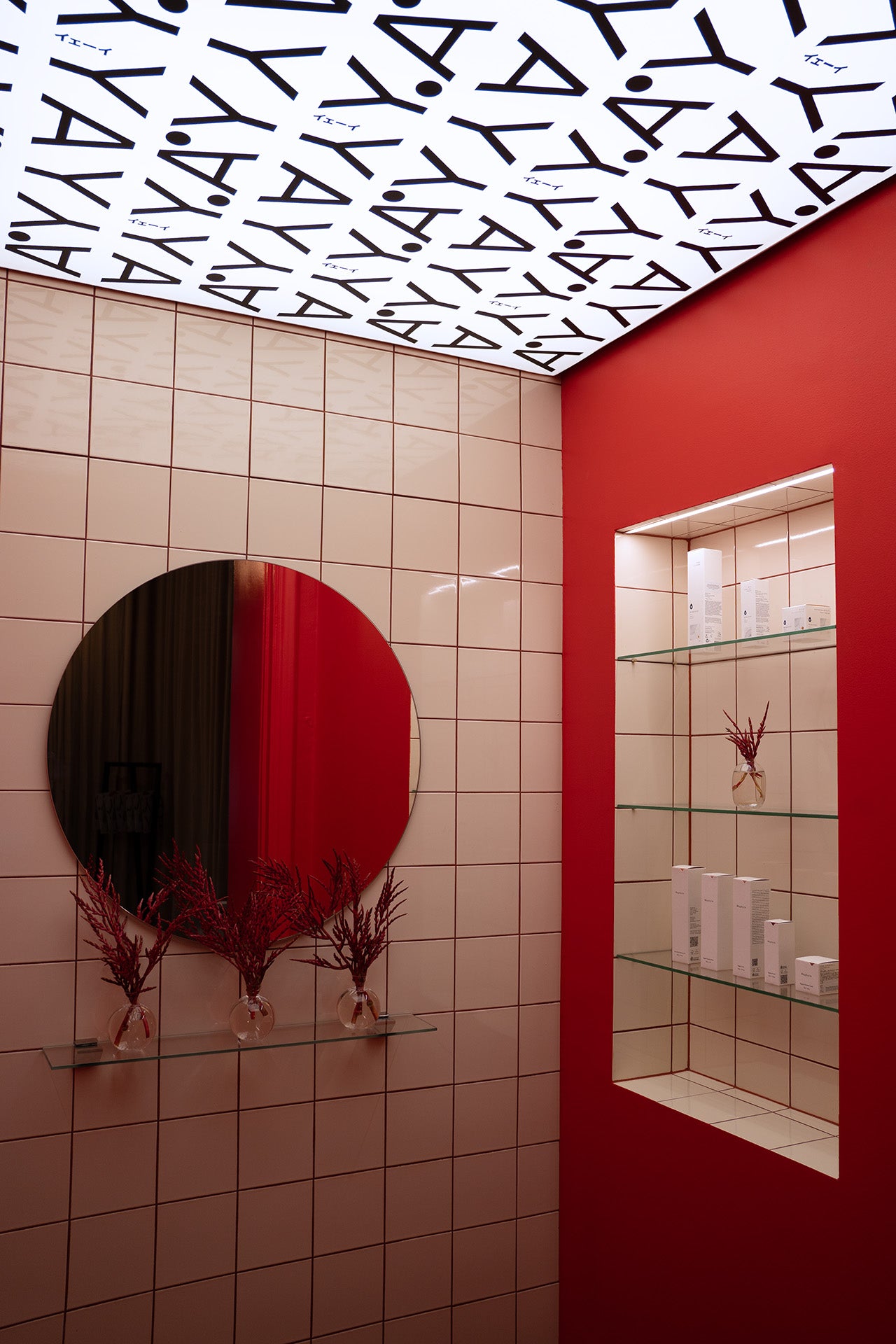 A stylish interior features red and beige tiled walls, a round mirror, and minimalist glass shelves displaying skincare products. The ceiling design repeats the word “YAY,” adding a playful contrast to the clean, contemporary aesthetic.