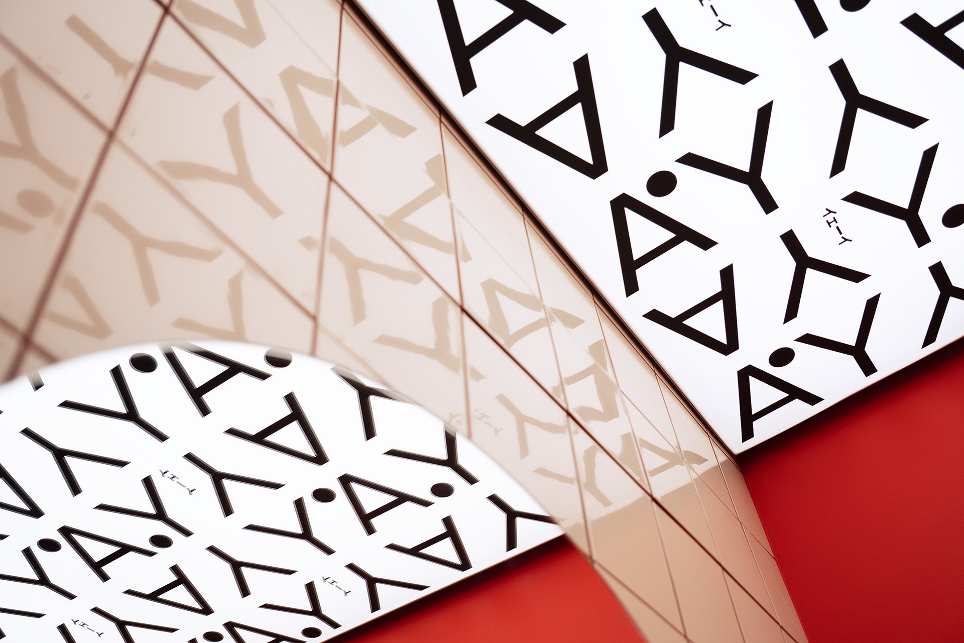 A close-up of a modern interior ceiling featuring a bold black “YAY” pattern on a white background, reflected in glossy tiled walls. The angled composition and red wall accents create a dynamic, graphic visual effect.