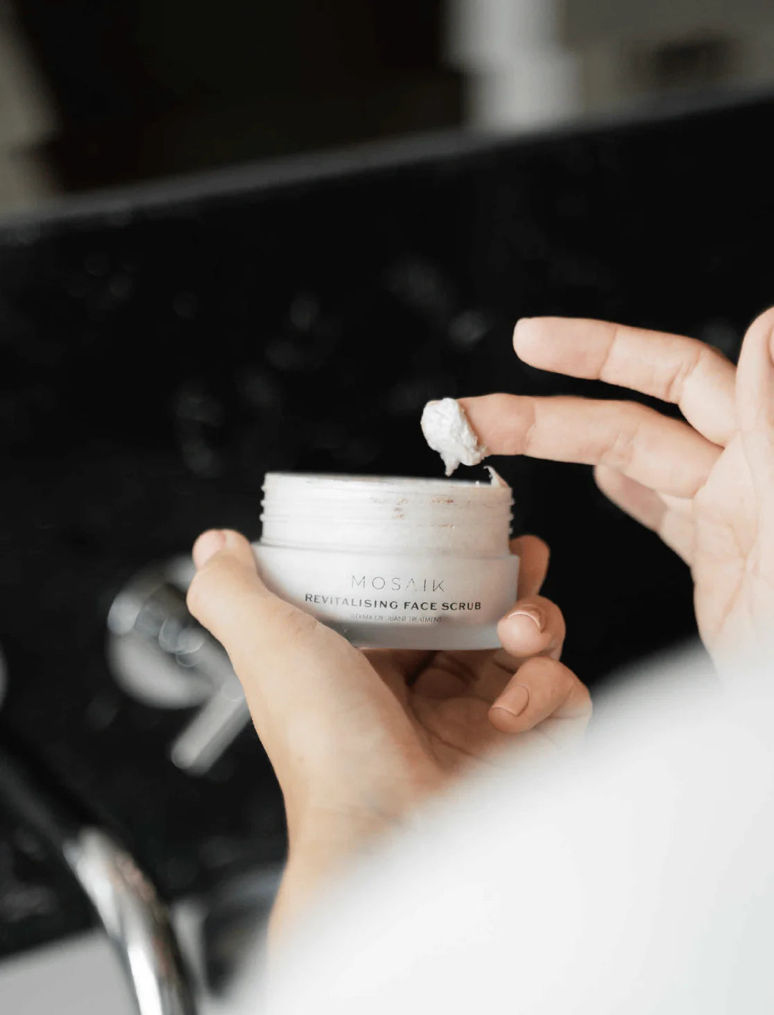 A person scoops a small amount of Mosaik Revitalising Face Scrub from its jar, showing the product’s creamy texture. The close-up shot highlights the luxurious consistency and gentle exfoliating formula.