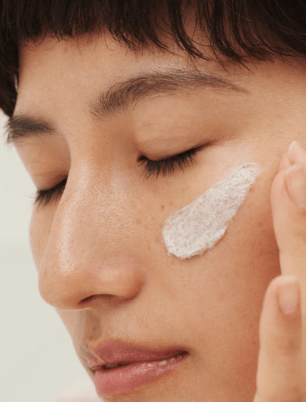 A person gently applies Mosaik Revitalising Face Scrub to their cheek, showing the exfoliating texture with fine natural particles. The close-up highlights smooth, hydrated skin and a calm self-care moment.