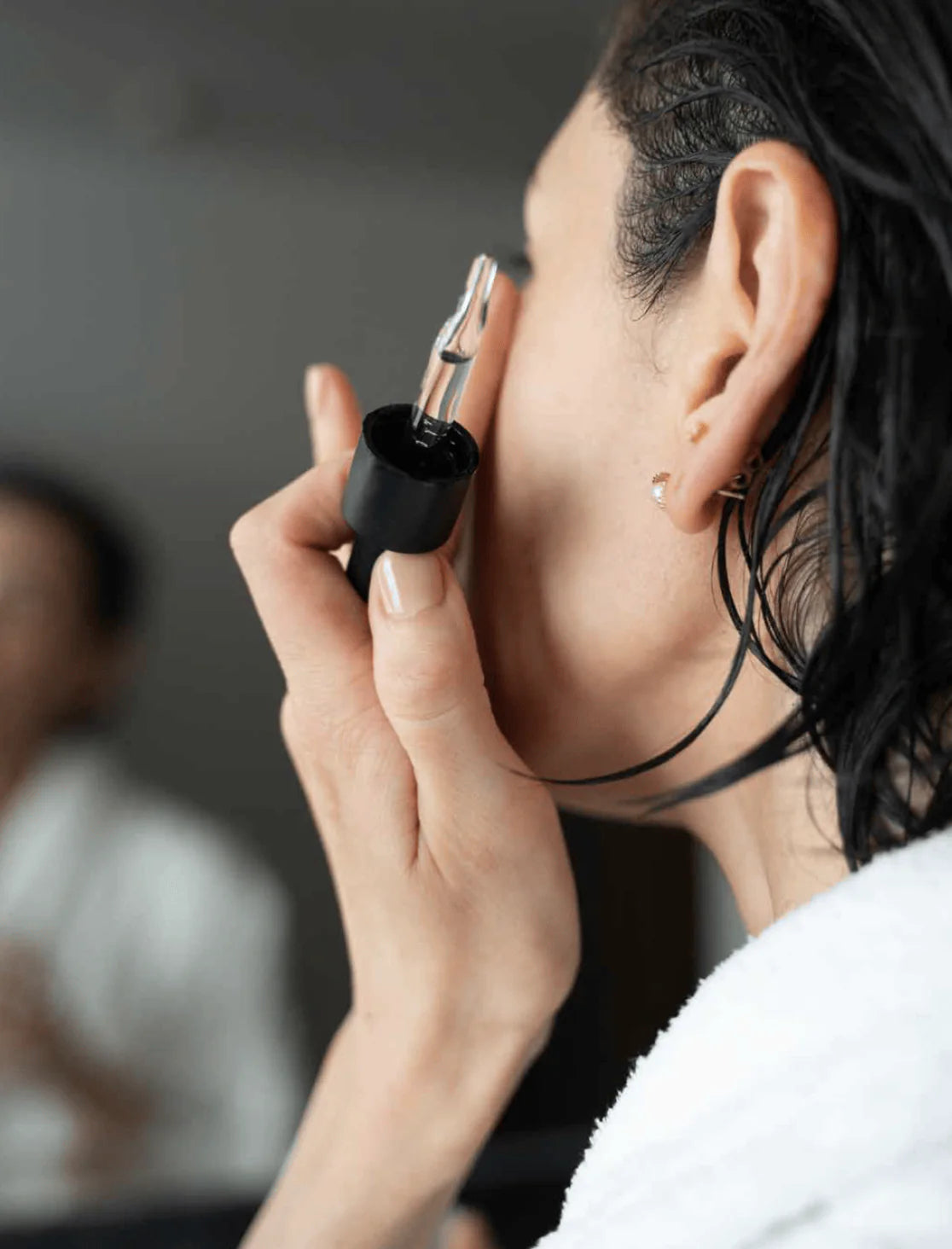 Close-up of a woman applying facial serum with a dropper to her skin after a shower, wearing a white robe, with a blurred bathroom mirror in the background.