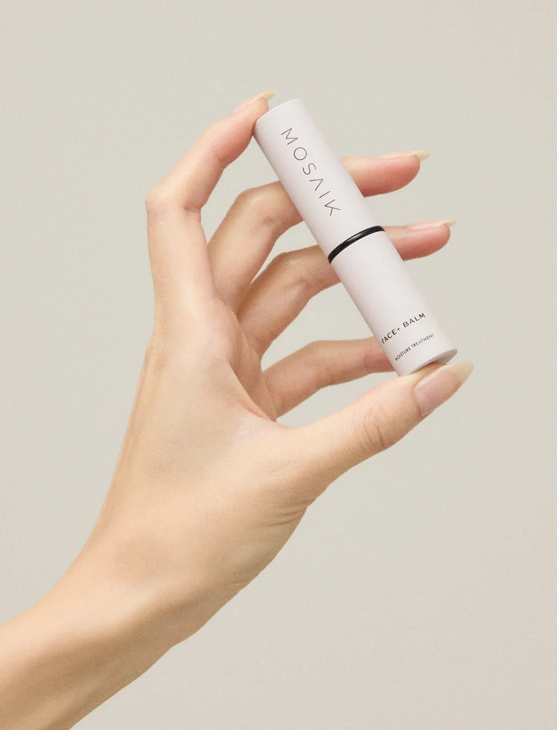 A hand delicately holds a tube of Mosaik Face Balm against a neutral background. The sleek, minimalist design with black text on a white tube reflects a refined and modern aesthetic.