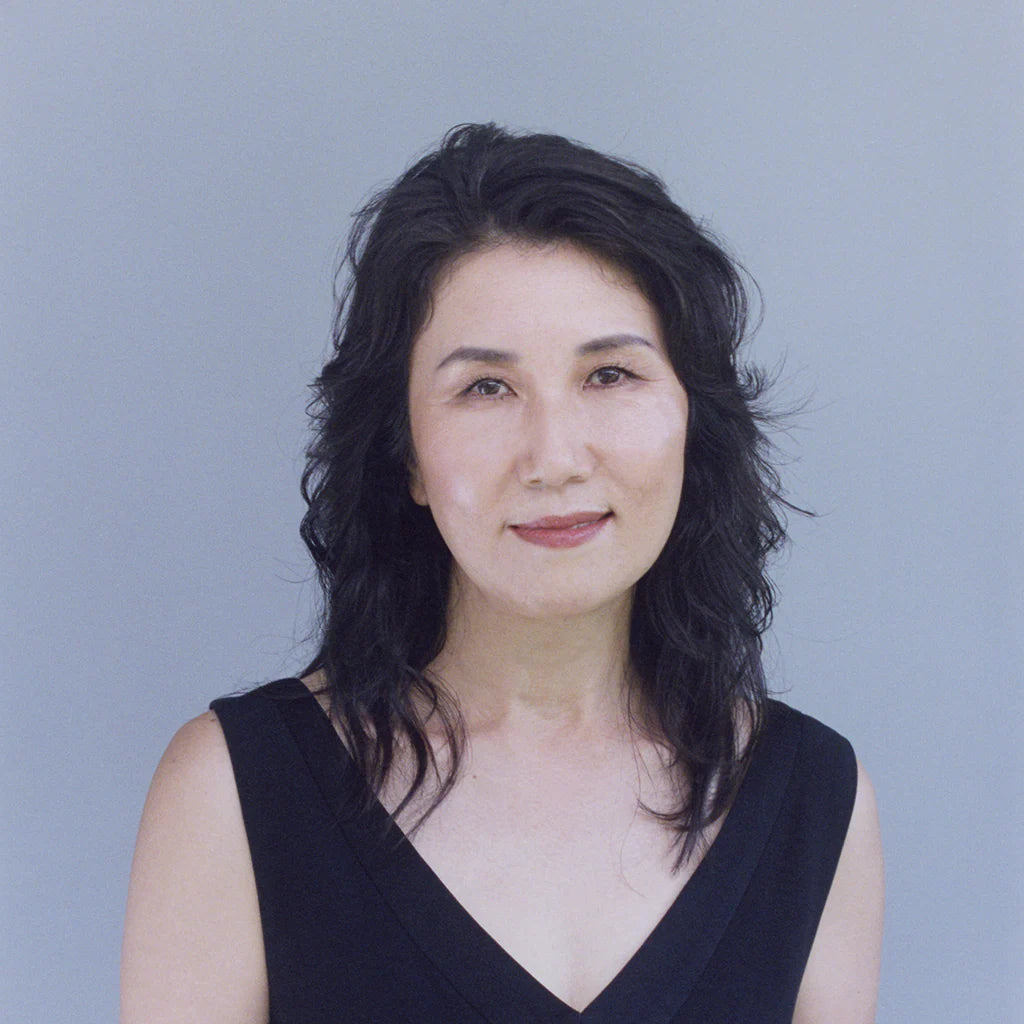 A person with medium-length dark hair and a calm expression stands against a light gray background. They are wearing a sleeveless black top and looking directly at the camera.