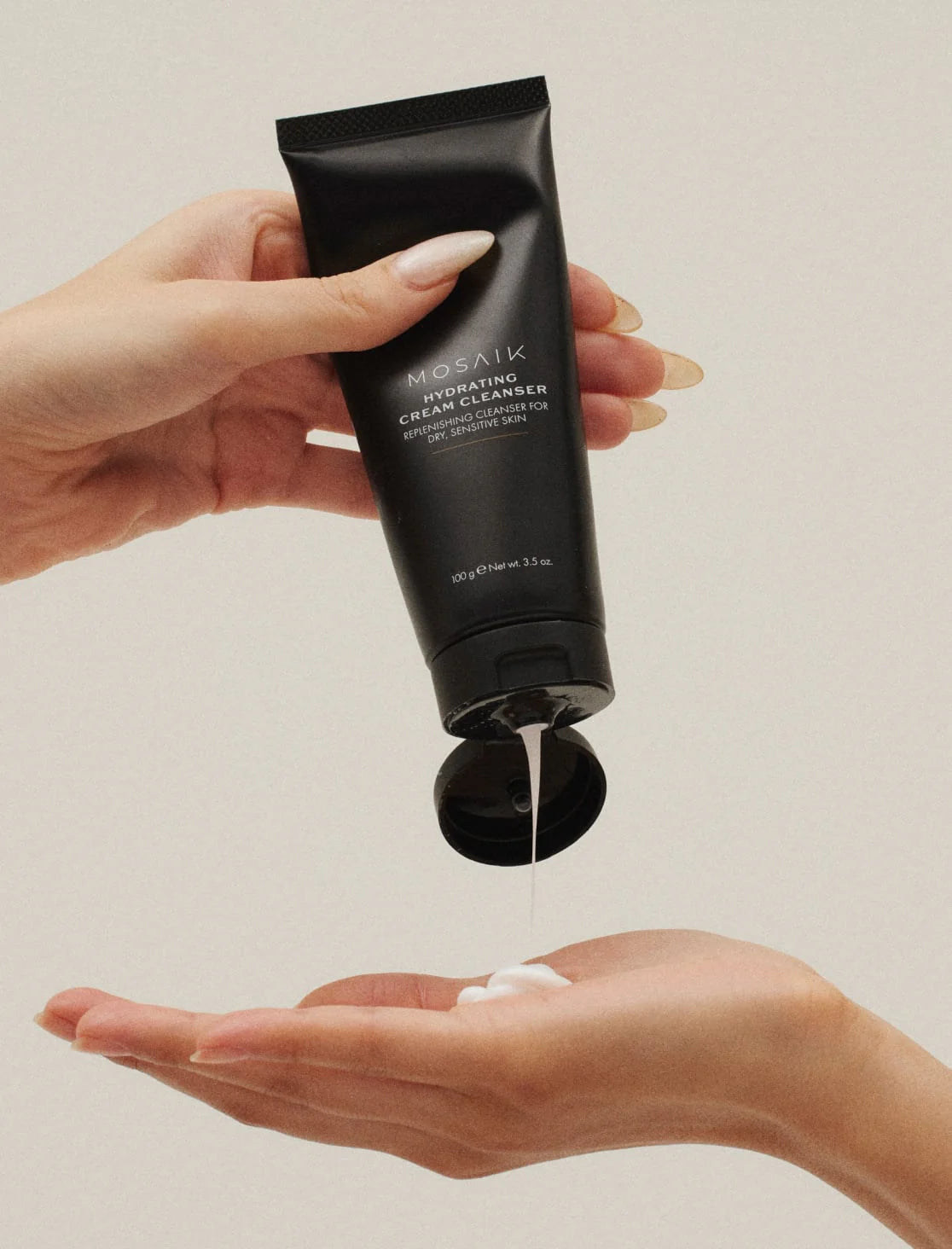 A person dispenses Mosaik Hydrating Cream Cleanser from a sleek black tube into their hand, showing its creamy texture. The minimalist scene highlights the product’s luxurious and gentle formula for dry, sensitive skin.