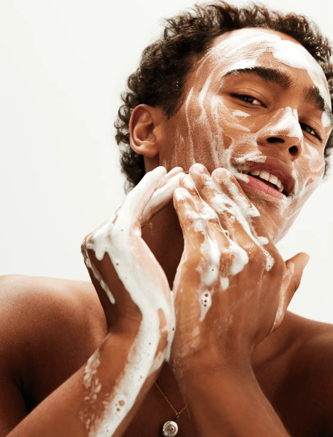 A person gently massages Mosaik Hydrating Cream Cleanser onto their face, creating a soft lather. The image captures a fresh, radiant moment of cleansing and self-care.