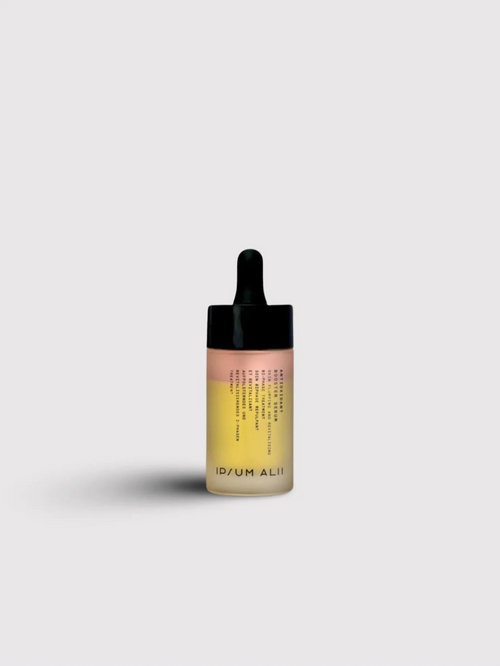 A bottle of Ipsum Ali facial oil with a black dropper cap standing against a plain light background. The bottle contains two visible layers of oil in pink and yellow tones.