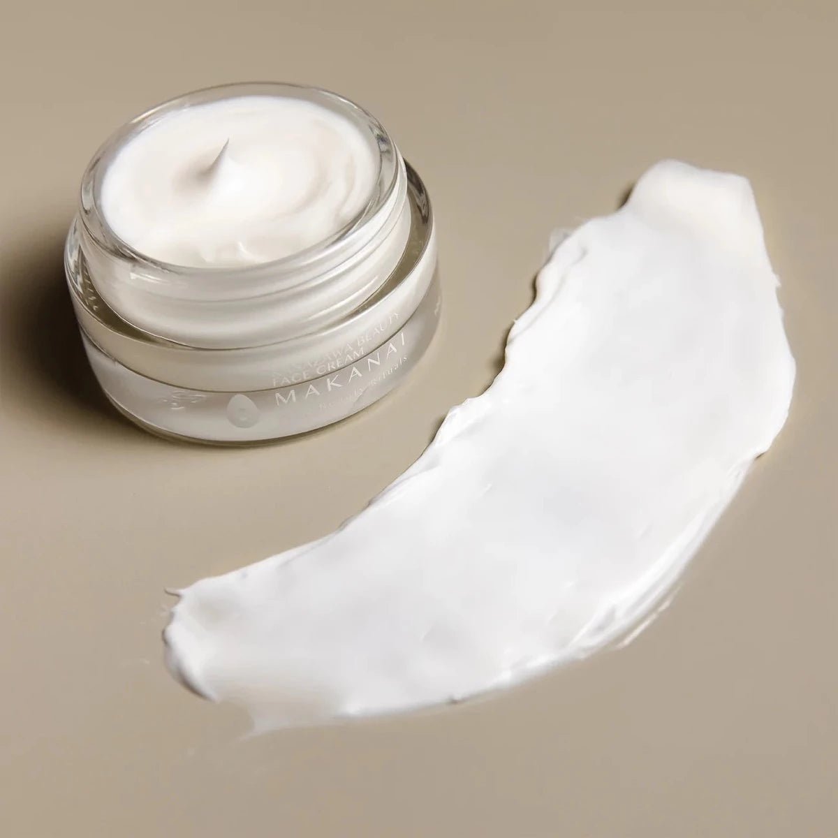 An open jar of Makanai Face Cream placed on a beige surface with a smooth swipe of cream next to it. The product appears rich and glossy, showcasing its creamy texture.