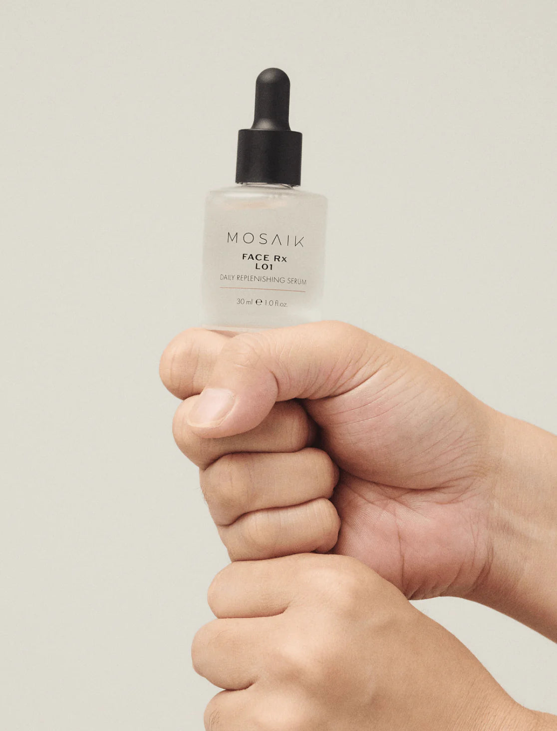Close-up of hands holding a bottle of MOSAIK Face Rx L01 Daily Replenishing Serum with a black dropper cap, against a soft neutral background.