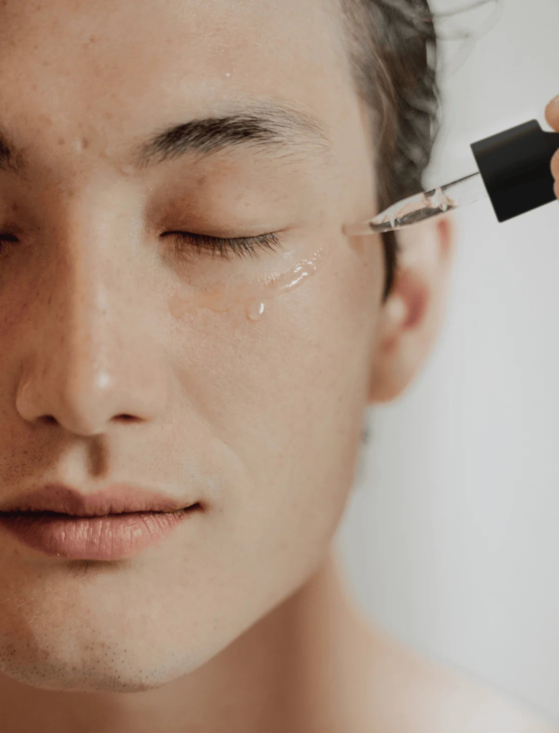 Close-up of a person applying MOSAIK Face Rx serum with a dropper to the skin near the eye, showing a glistening layer of product for a clean and hydrated finish.