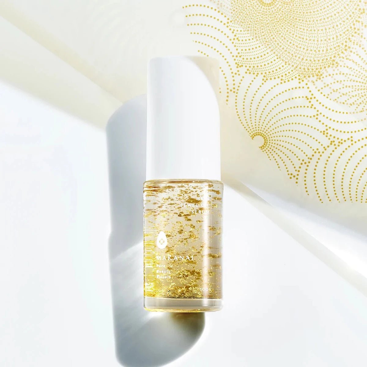 A bottle of Makanai Skin Jewel Serum filled with clear liquid and visible gold flakes, shown on a light background with a soft shadow. The design includes a white cap and subtle gold pattern detailing in the backdrop.
