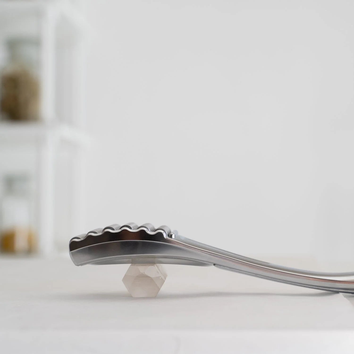 Slim Cera silver facial roller resting on a crystal base, side view showing its curved ergonomic design and textured roller head, placed on a white surface with a soft background.