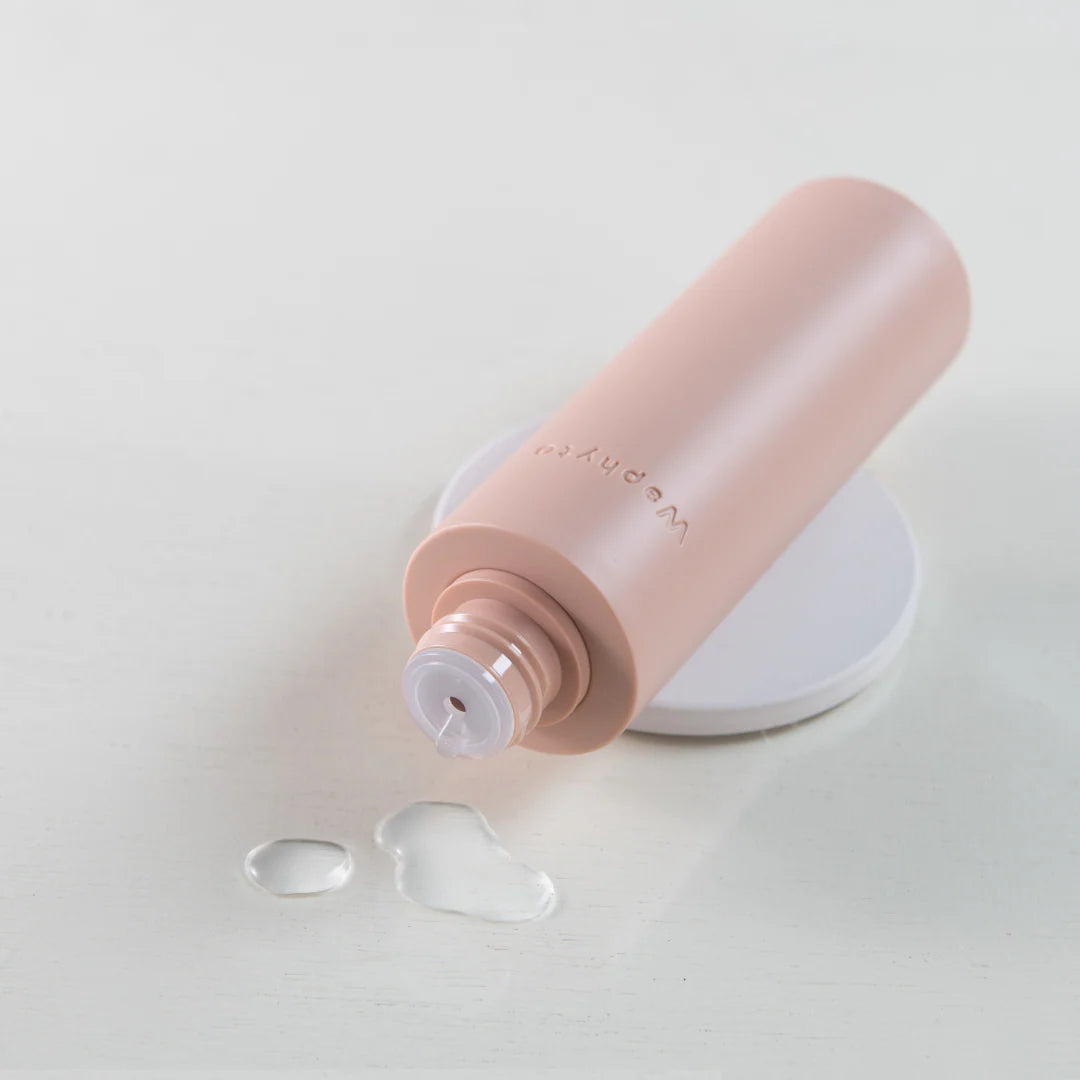 A light pink Waphyto bottle lies on its side with a few drops of liquid beside the opening. The image highlights the product’s clean, minimal design and fresh texture.