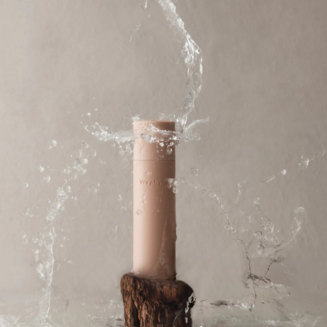 A beige Waphyto bottle stands on a piece of wood as water splashes dramatically around it. The image conveys freshness, purity, and a connection to natural elements.